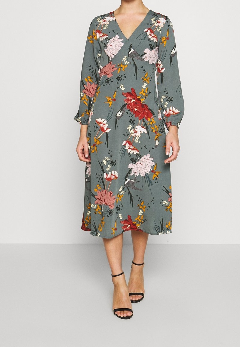 Mujer vistiendo un vestido verde de longitud media con un patrón floral en rojo, rosa, amarillo y blanco, combinado con tacones negros de pulsera al tobillo.