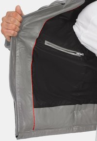 Grijze leren jasinterieur met een zwarte voering, een ritsvak en rode accenten langs de naden. Ritsafsluiting zichtbaar.