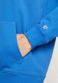 Blå sweatshirt med texturerad tyg, framficka och ribbade muddar. Har en liten röd och vit logotyp på ärmen.