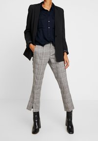 Černá sako přes námořnickou košili s knoflíky, doplněné šedými kostkovanými kalhotami ve slim fit s bočními rozparky a černými botami na podpatku.