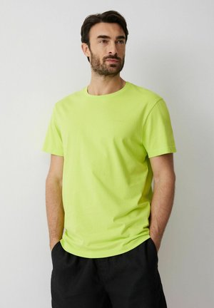 Mężczyzna o ciemnych włosach i brodzie, ubrany w limonkowo-zielony t-shirt z krótkim rękawem i czarne spodnie, stojący z rękami w kieszeniach na tle gładkiej ściany.