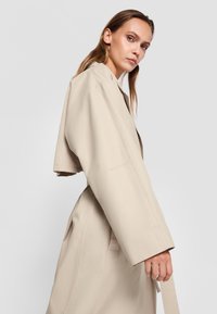 Beige trench coat with a wide lapel, cropped sleeves, and a belted waist. Smooth texture with side pockets, shown at a slight angle.