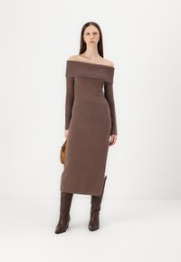 Ruda off-the-shoulder mezginė su ilgomis rankovėmis, susiraukšlėtu apykaklės kraštu, prigludusia forma ir kelių ilgio sijonu, derinama su rudais aukštakulniais batais.
