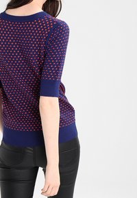 Svetr s krátkým rukávem v námořnické modré barvě s červeným trojúhelníkovým vzorem, žebrovaný výstřih a lem, přiléhavý střih, kombinovaný s černými texturovanými kalhotami.