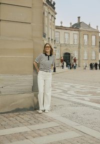 Gestreepte zwart-witte polo met een zwarte kraag, witte wijde broek en witte sneakers. Gele zonnebril accentueert de outfit.