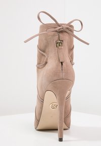 Beige ruskind ankelstøvle med høj hæl og snørebånd, baglås og hardware i guldfarve. Glat tekstur og formet silhuet.