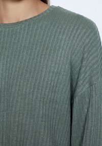 Pull vert en maille côtelée avec un col rond, présentant une texture douce et une coupe décontractée, montrant un gros plan des détails du tissu.
