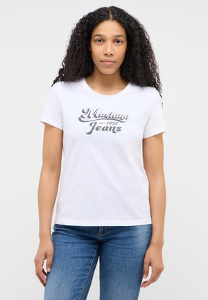 Frau mit lockigem Haar, die ein weißes "Mustang Jeans since 1932" T-Shirt und blaue Jeans trägt und vor einem schlichten hellen Hintergrund steht.