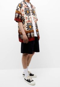 Chemise à manches courtes avec un motif floral en jaune, marron et noir sur un fond crème ; portée avec un short foncé et des baskets.