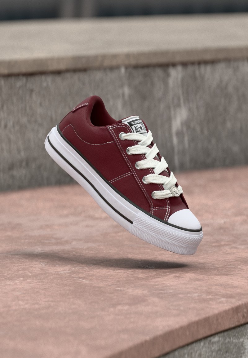 Μπορντό χαμηλά παπούτσια Converse με άσπρα κορδόνια, λαστιχένια μύτη και λογότυπο στη γλώσσα, αιωρούμενα πάνω από μια πέτρινη επιφάνεια.