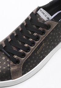 Brązowe i czarne materiałowe trampki z wzorem gwiazdy, czarnymi sznurowadłami, białą gumową podeszwą i metalową ozdobą logo przy sznurowadłach.