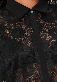 Crna bluza od čipke s cvjetnim uzorkom i čvrstim crnim ovratnikom. Uključuje ukrasne gumbe. Prozirna tekstura s detaljnim dizajnom.