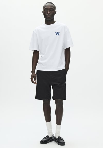 Λευκό υπερμεγέθες t-shirt με ναυτικό λογότυπο, σε συνδυασμό με μαύρο σορτς. Το μοντέλο φοράει λευκές κάλτσες και μαύρα μοκασίνια, στέκεται μπροστά σε απλό φόντο.