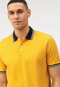 Geel poloshirt met een zwarte kraag voorzien van witte strepen. Twee knopen bij de kraag, textiel met structuur, normale pasvorm. Logo op de borst geplaatst.