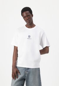 Camiseta de algodón blanca con mangas cortas y cuello redondo, con un logo en azul marino que lleva el texto "ORIGINALS" centrado en el pecho.