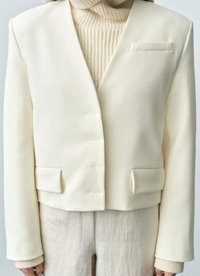 Elfenbensfärgad blazer med en strukturerad design, framknappar och två fickor med klaff. Tyget verkar slätt med en något matt yta; utan krage.