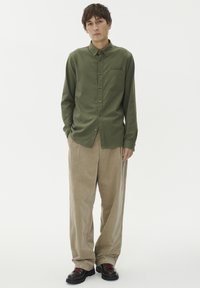 Olivnozeleni srajčni zgornji del z žepom na prsih, v kombinaciji z ohlapnimi bež kordnimi hlačami in temnimi čevlji z rdečimi vezalkami.