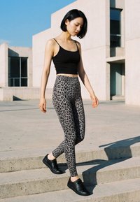 Femeie cu păr scurt și negru purtând un top scurt negru și colanți cu imprimeu leopard, coborând scări din beton în aer liber, sub lumina puternică a soarelui.