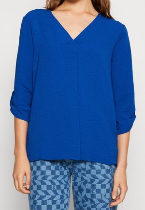 Frau trägt eine leuchtend blaue Bluse mit V-Ausschnitt und Dreiviertelärmeln sowie blau gemusterte karierte Hose.