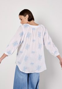 Vit blus med ljusblåt blommönster, samlade detaljer bak och lösa puffärmar. Ytterligare denimbyxor syns nedanför.