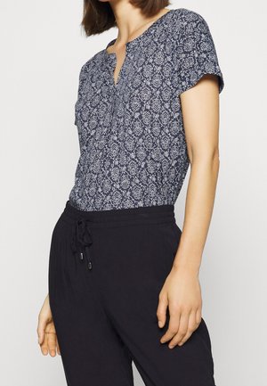 Nainen, joka pukeutunut lyhythihaiseen tummansiniseen puseroon, jossa on valkoisia kukkakuvioita, ja mustiin nyörillä kiristettäviin housuihin. Kuvassa näkyy kaulasta lantioihin.