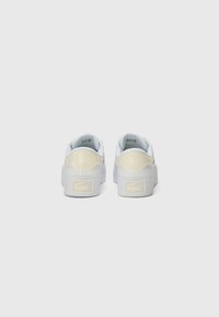 Hvite sneakers med en glatt læroverdel, kontrasterende lys gule detaljer og teksturerte logoer på hælen og tungen. Flat gummisåle.