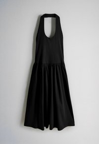 Robe midi noire sans manches avec col licou et jupe froncée suspendue à un mur gris clair.