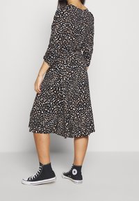 Frau in einem schwarzen knielangen Kleid mit weißen und braunen Flecken, dreiviertel Ärmeln, kombiniert mit schwarzen High-Top Converse Sneakers.