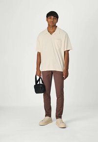 Helles beiges Kurzarm-Polohemd mit Tasche, braune Slim-Fit-Hose, cremefarbige Sneaker und schwarze Umhängetasche, stehend vor einem hellen Hintergrund.