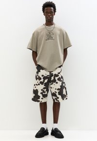 Světle béžové oversized tričko s grafickým potiskem, doplněné černobílými kraťasy s kravským vzorem. Celkový look dotvářejí černé mokasíny a bílé ponožky.