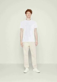 Young man with curly red hair wearing a plain white T-shirt, beige pants, and white sneakers, standing against a light neutral background.