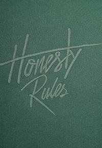 Grön textil med upphöjd, broderad text som läser "Ärlighet Råder." Slät textur med minimalistisk design och ett subtilt mönster.