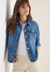 Femme aux longs cheveux bruns portant une veste en jean bleue sur un chemisier blanc et un pantalon beige, debout et appuyée contre un mur clair.