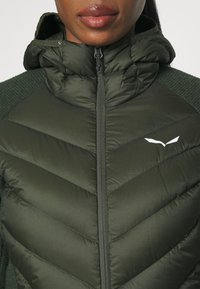 Puffy green jacket with a high collar, zip closure, and chevron quilting. Features knitted accents on the shoulders and a logo.