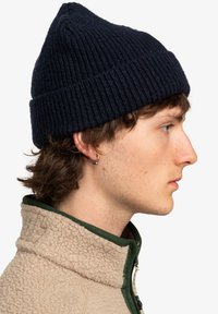 Gorro de punto navy con textura acanalada, presenta una parte superior puntiaguda y un dobladillo plegable. Se lleva con una chaqueta de forro polar beige con detalles en verde.