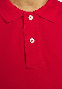 Červené polo tričko s vroubkovaným límcem a třemi bílými knoflíky, které mají červené prošívání. Tkanina má texturovanou, prodyšnou kvalitu.