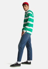 Długorękawowa koszulka polo w zielono-białe paski, niebieskie dżinsy, czarne buty z zielonymi akcentami oraz czerwona czapka. Model stoi bokiem.
