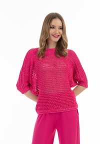 Różowy sweter dziergany o luźnym kroju, z krótkimi prążkowanymi rękawami i otwartą fakturą. W połączeniu z dopasowanymi różowymi spodniami.