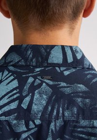 Camicia a maniche corte in tessuto blu scuro con motivi a foglia blu chiaro, colletto button-down e un piccolo etichetta metallica sul retro.