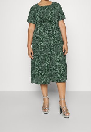 Mujer con un vestido verde hasta la rodilla con un estampado de hojas negras y sandalias plateadas de tacón alto, de pie frente a un fondo blanco liso.