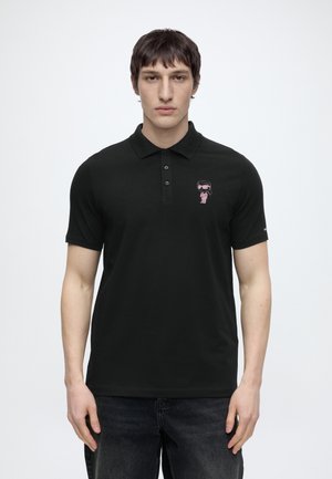 Mann trägt ein schwarzes kurzärmeliges Poloshirt mit einer kleinen pinkfarbenen Grafik einer stilisierten Figur auf der linken Brust.