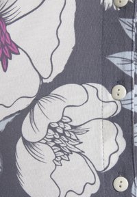 Blommönstrad tyg med stora vita och lila blommor på en grå bakgrund. Inkluderar ljusa knappar och synliga sömnadsdetaljer.