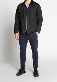 Černá látková bunda s manšestrovým límcem, která má dvě náprsní kapsy a dvě boční kapsy. Nahoře je nošena šedá košile a tmavě modré kalhoty, s černými botami.