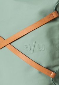 Grüne Stofftasche mit texturierter Oberfläche, ausgestattet mit einem hellbraunen Ledertrageriemen und geprägt mit dem Logo "a/u." Das gekreuzte Riemendesign betont die Form.