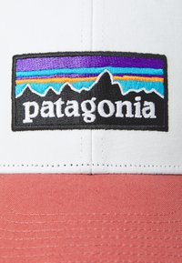 Bela in koralna bejzbolska kapa z črnim vezenim logotipom Patagonia, ki vsebuje večbarvno gorsko in črtno zasnovo.