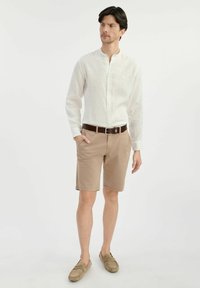 Biała lniana koszula z kołnierzykiem mandaryńskim, beżowe szorty, brązowy skórzany pasek i brązowe loafersy, stojący na jednolitym tle.