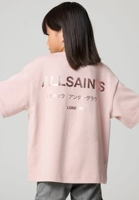 Vaaleanpunainen oversize T-paita, joka on valmistettu puuvillasta. T-paidan selässä on valkoinen ja metallinen printti, jossa lukee "ALLSAINTS" japanin merkeillä ja "LONDON".