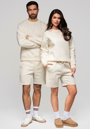 Sudadera de color crema y pantalones cortos, hechos de una tela suave. La sudadera tiene un escote redondo; los pantalones cortos cuentan con una cinturilla elástica.