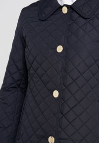 Marinblå quiltad jacka med stor krage, diagonal sömnad, slät textur och två stora guldfärgade knappar framtill.