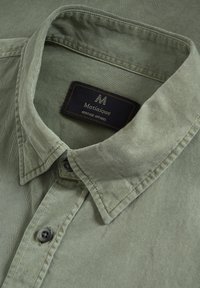 Sõlmikuga oliiviroheline triiksärk hallide nuppudega ja sees must silt tekstiga "Matinique Heritage Apparel".